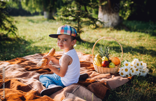 The lovely little boy in baseball cap sitting on a plaid and eating traditional french baguette. Summer day. Child on plaid eatin bread. Аdvertising produstion. Near pine forest and lake.  photo