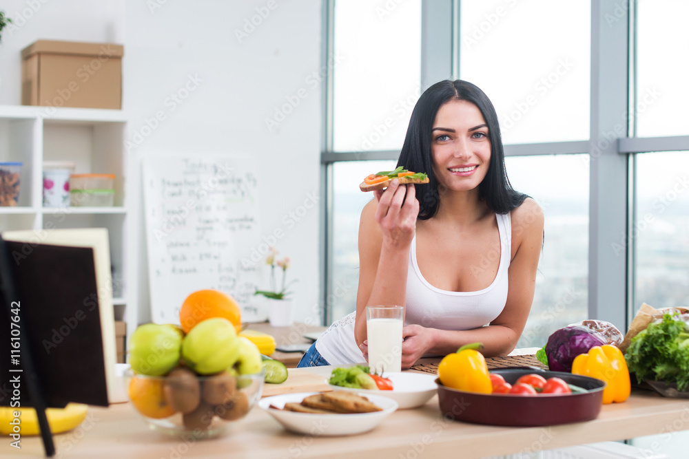 Young female standing in kitchen leaning at wooden counter, having sandwich and a glass of milk for lunch. Young female standing in kitchen leaning at wooden counter, having sandwich and a glass of milk for lunch.