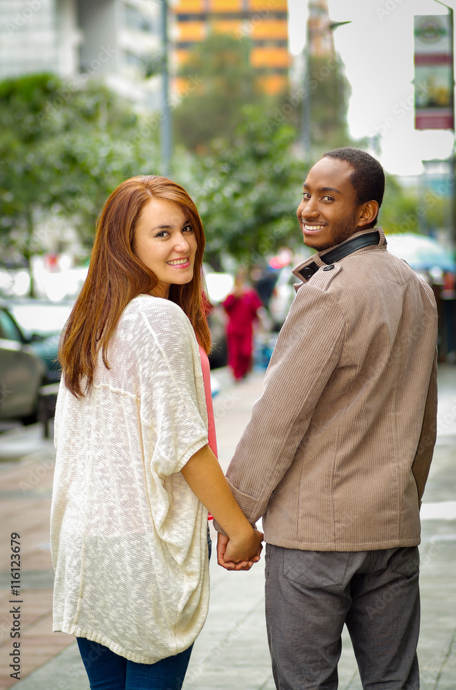 Interracial happy charming couple wearing casual clothes walking holding hands, turns around heads for camera in outdoors urban environment