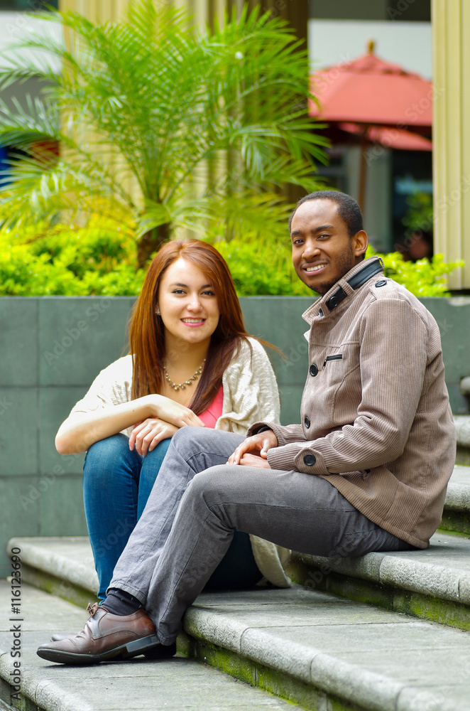Interracial happy charming couple sitting on steps in front of building interacting and smiling for camera Interracial happy charming couple sitting on steps in front of building interacting and smiling for camera