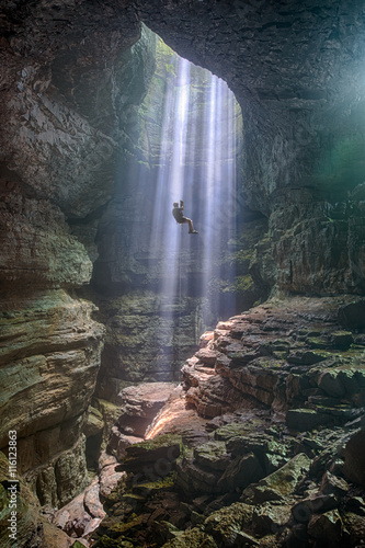 The mist in a dark wet cave is naturally lit by shafts of light coming through an opening in the ceiling.  Using a long rope a man rappels into the mist bathed by sunlight