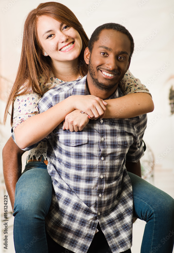 Interracial couple wearing casual clothes interacting having fun, man carrying woman on his back, both smiling happily, white studio background Interracial couple wearing casual clothes interacting having fun, man carrying woman on his back, both smiling happily, white studio background