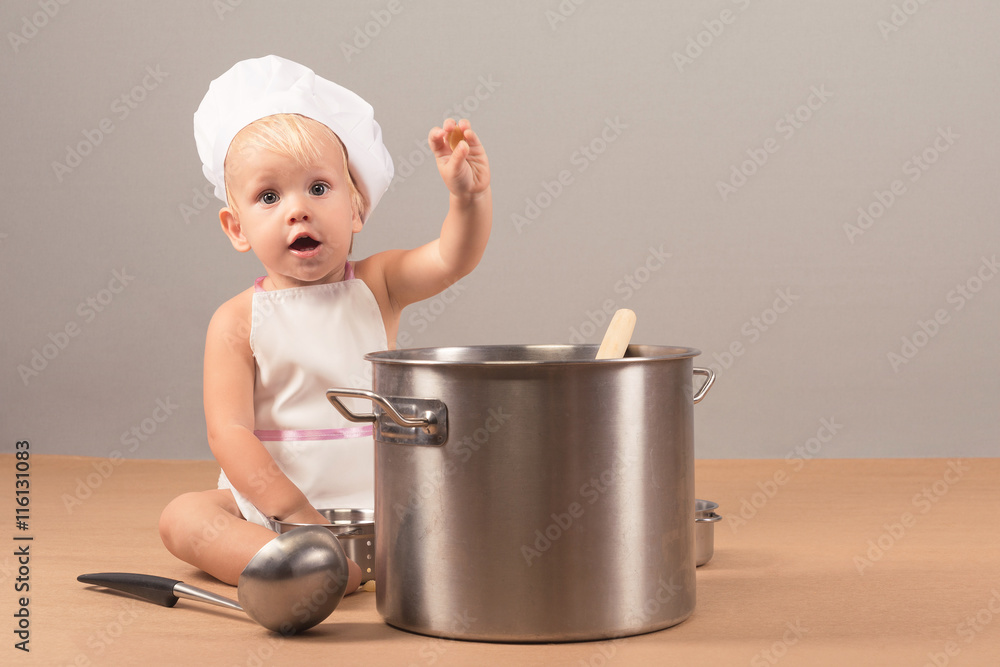 Charming and surprised kid in an apron and chef hat sitting next to a big pot of soup and holding something in his hand Charming and surprised kid in an apron and chef hat sitting next to a big pot of soup and holding something in his hand