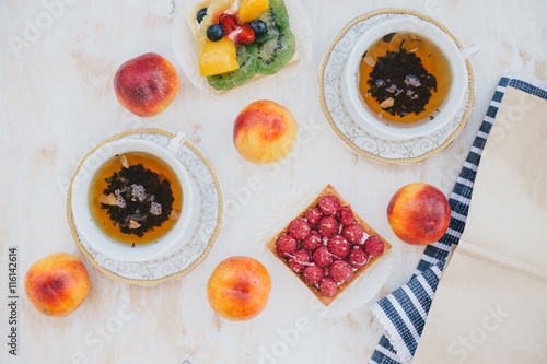 Still life with two cups of tea in a vintage cups and  two tarts with fresh fruits: raspberries, blueberries, kiwi, strawberries and peaches on a white vintage background