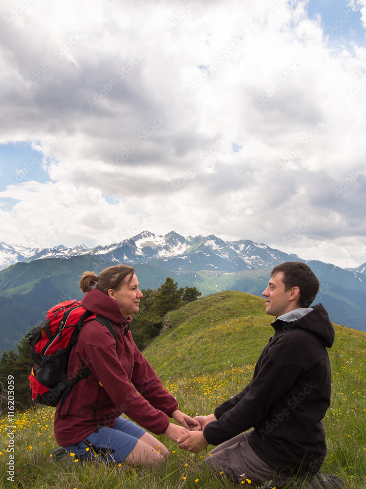 Two young people sitting on the grass and holding hands on a background of mountains with clouds