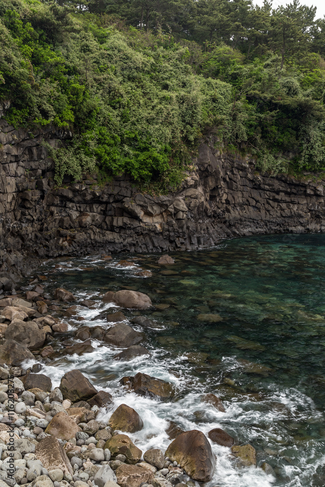 Obraz premium Rocky coastline and steep cliff next to Sojeongbang Falls on Jeju Island in South Korea.