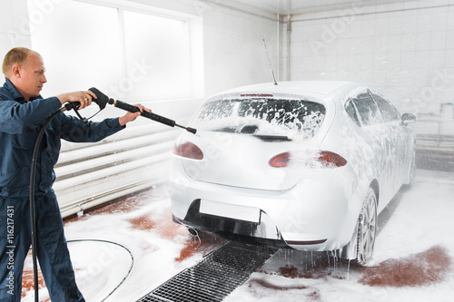 Handle carwash concept, man with hose wash white car at service station. Garage worker washing automobile with nosepiece photo