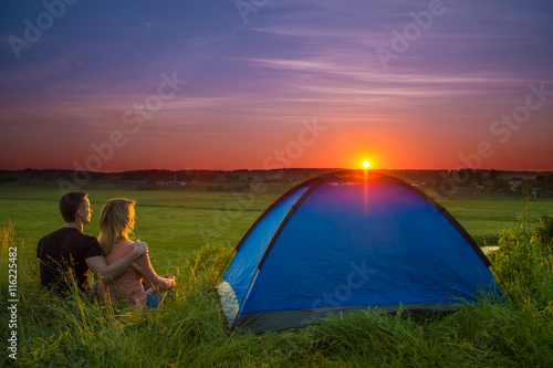 The couple sit near camping tent by sunset (sunrise) background. Wide anlge