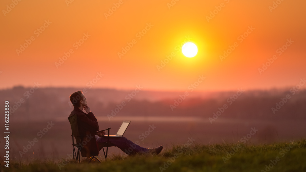 The man sit and work on the laptop by sunset (sunrise) background on the top of the hill The man sit and work on the laptop by sunset (sunrise) background on the top of the hill