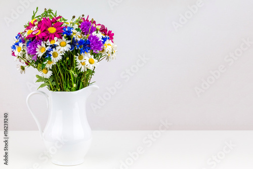 Wildflowers in white ceramic jug with copy space. Wild flower bouquet on white table. Bunch of wild herbs and flowers in a white jug. Wild flowers in a vase.