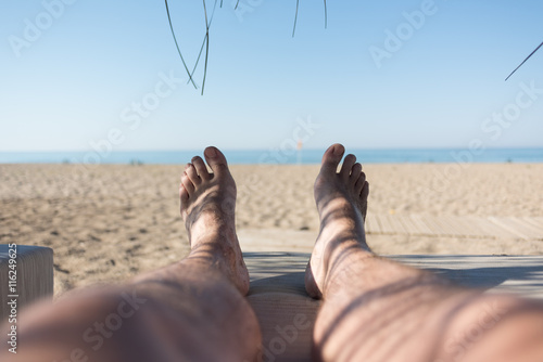 Fototapeta Naklejka Na Ścianę i Meble -  Male feet on lounge first person view from bungalow on the sea with sand beach