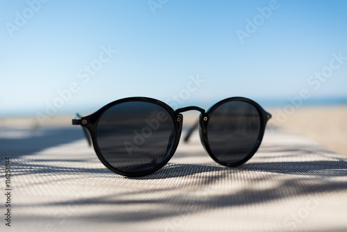Fototapeta Naklejka Na Ścianę i Meble -  Female sunglasses on a sun lounger with defocused sand beach and sea on the background