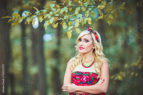 Girl in red dress standing on the background of trees in the forest. Woman in red dres and bandana. Dress painted flowers. Model in summer meadow forest\park. Advertising photoshoot. photo