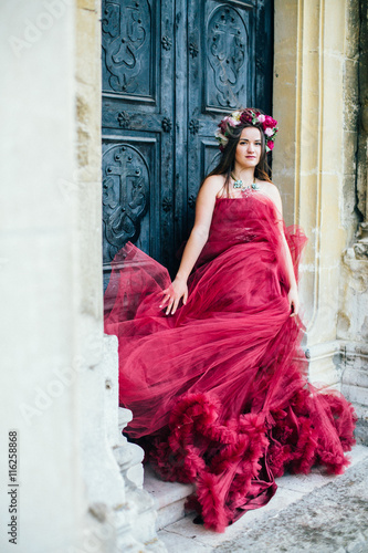 Beautiful young woman in a bright red dress and a crown of white spring flowers posing against the backdrop of the ancient door. Wind fanning,overblow, fan cloud dress. Brunet girl  with wrath.  © nataliakabliuk