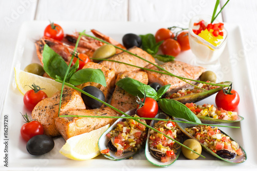 Close-up of plate with assorted cooked seafood and spicy sauce on white wooden background. Marine cuisine