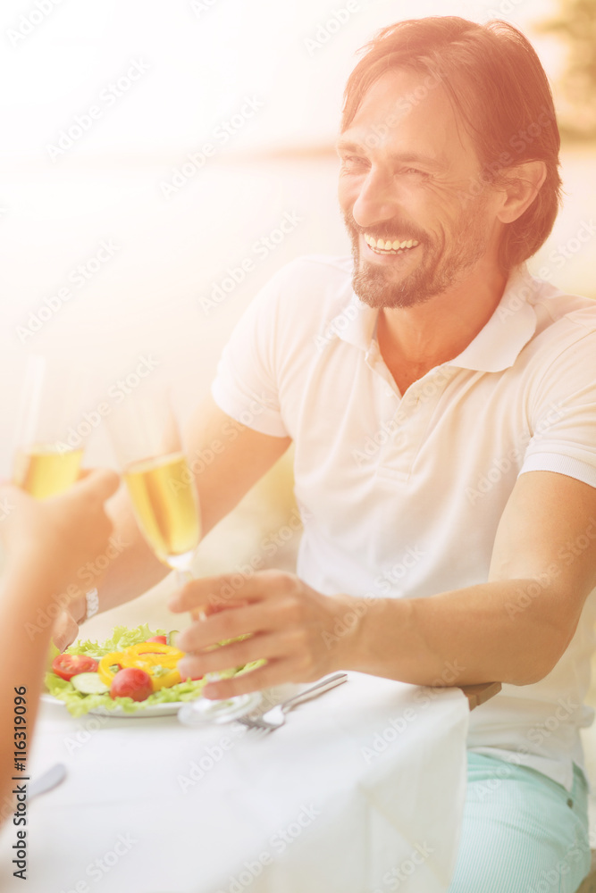 Closeup toned picture of happy man toothy smiling while sitting at table in restaurant or cafe by sea. Handsome man holding glass of expensive wine. Closeup toned picture of happy man toothy smiling while sitting at table in restaurant or cafe by sea. Handsome man holding glass of expensive wine.