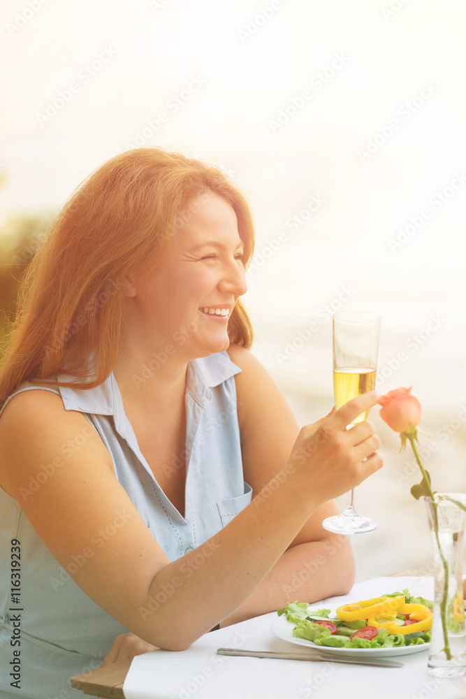 Closeup toned profile of beautiful red-haired mature woman smiling and holding glass of wine. Pretty woman looking away while spending time in restaurant or cafe by sea.