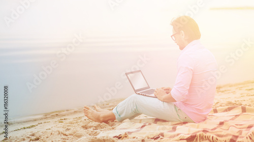Toned picture of handsome mature freelancer working on laptop computer. Author in glasses writing poem at beach. He is becoming inspired by beautiful sea.