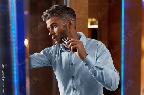 Man With Electric Razor Shaving Face In Bathroom. Men Grooming
