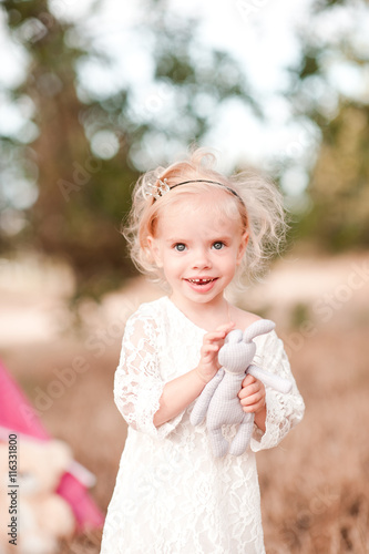 Smiling baby girl 1-2 year old holding toy rabbit outdoors. Looking at camera. Childhood. Happiness.