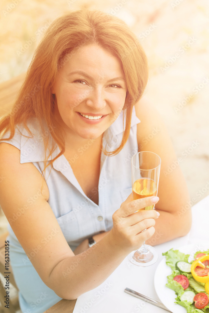 Closeup toned image of beautiful woman holding glass of white wine and smiling to camera. Red-haired woman in blue dress sitting in restaurant or cafe. Closeup toned image of beautiful woman holding glass of white wine and smiling to camera. Red-haired woman in blue dress sitting in restaurant or cafe.