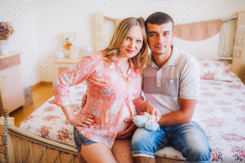 Image of happy future dad holding baby shoes on the belly of his caucasian pregnant wife lying on bed at home.