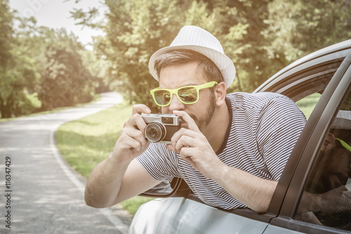 Portrait of young excited tourist guy taking photos through the window from driver’s seat on road trip travel vacation by using vintage retro camera.