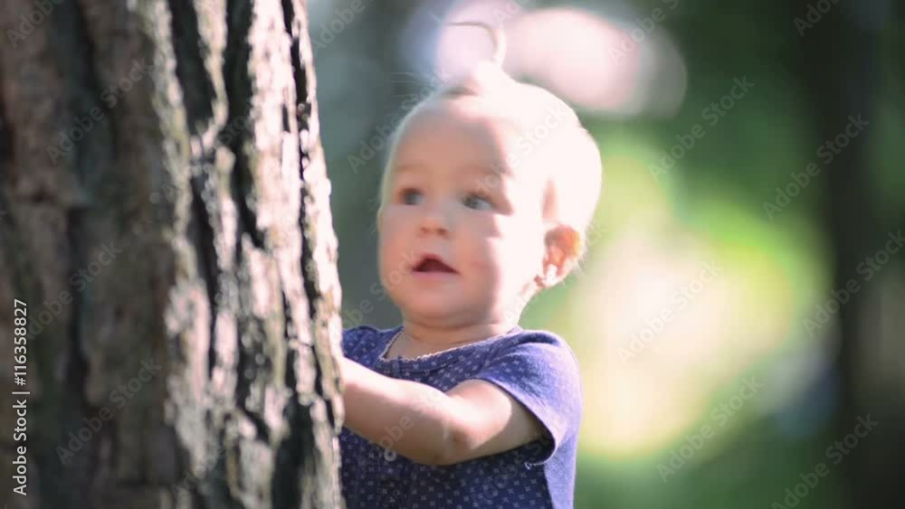 Baby girl one year old playing hide and seek, hiding behind a big tree, close-up Baby girl one year old playing hide and seek, hiding behind a big tree, close-up
