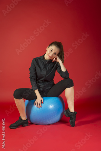 Portrait of a sporty woman, aerobic instructor sitting on a exercise ball, studio shot on red background