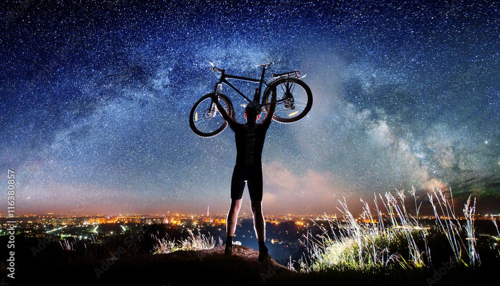 Obraz premium Silhouette of biker hold his bicycle above in the hands on the top of the mountain nearly city. Milky way and night city shining on the background