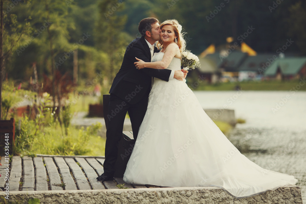 Bride holds groom's hand while he hugs her on the lake's shore Bride holds groom's hand while he hugs her on the lake's shore
