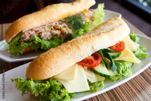 Cheese and salad baguette on white plate. Tuna baguette in backg