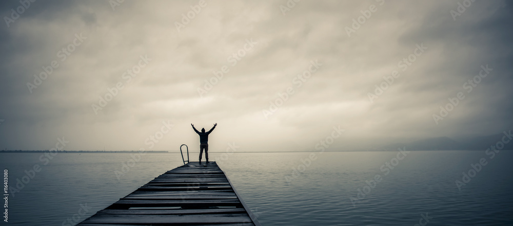 Man standing on a old wooden dock with raised hands - obrazy, fototapety, plakaty 