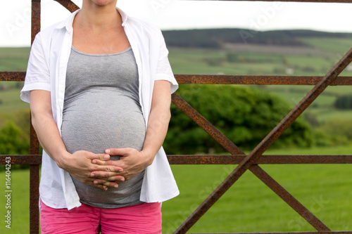 pregnant woman  standing in front of rusty fence in front of a filed.