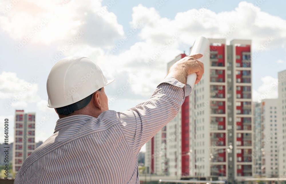 An engineer in white helmet pointing with blueprints in his hand on buildings on a background An engineer in white helmet pointing with blueprints in his hand on buildings on a background