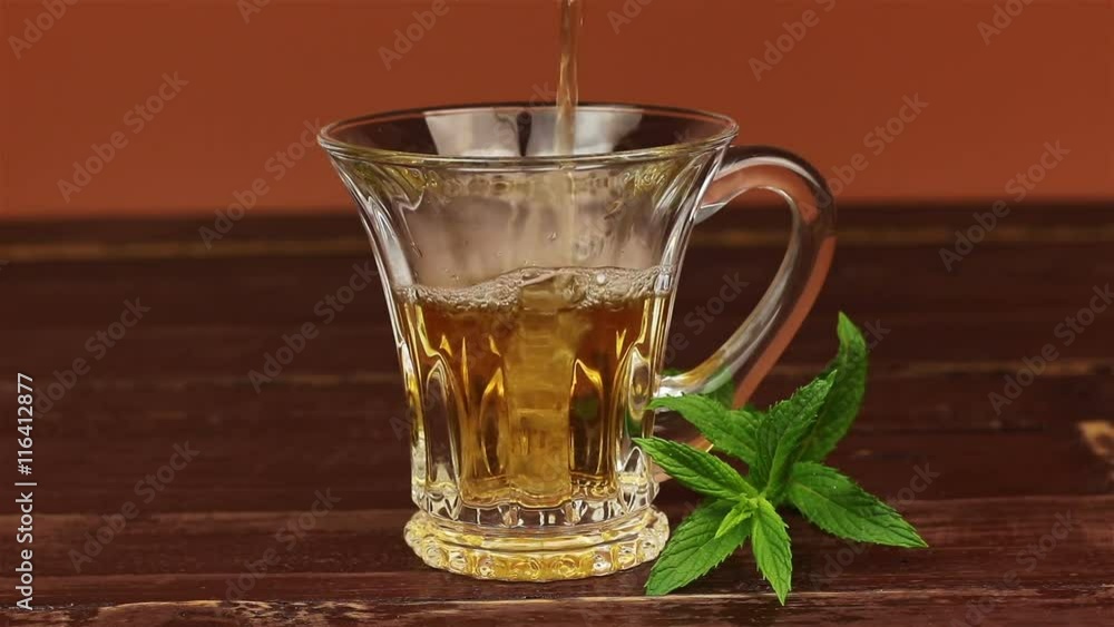 Tea being poured into glass tea cup and mint leaves on brown wooden table Tea being poured into glass tea cup and mint leaves on brown wooden table