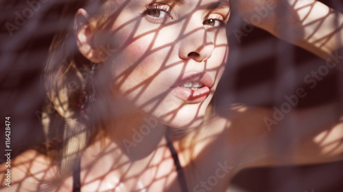 Closeup portrait of beautiful blonde woman wearing black swimwear sitting on a rooftop in a sunny summer day with shadow net reflection from metal sunshade on her face over black wall