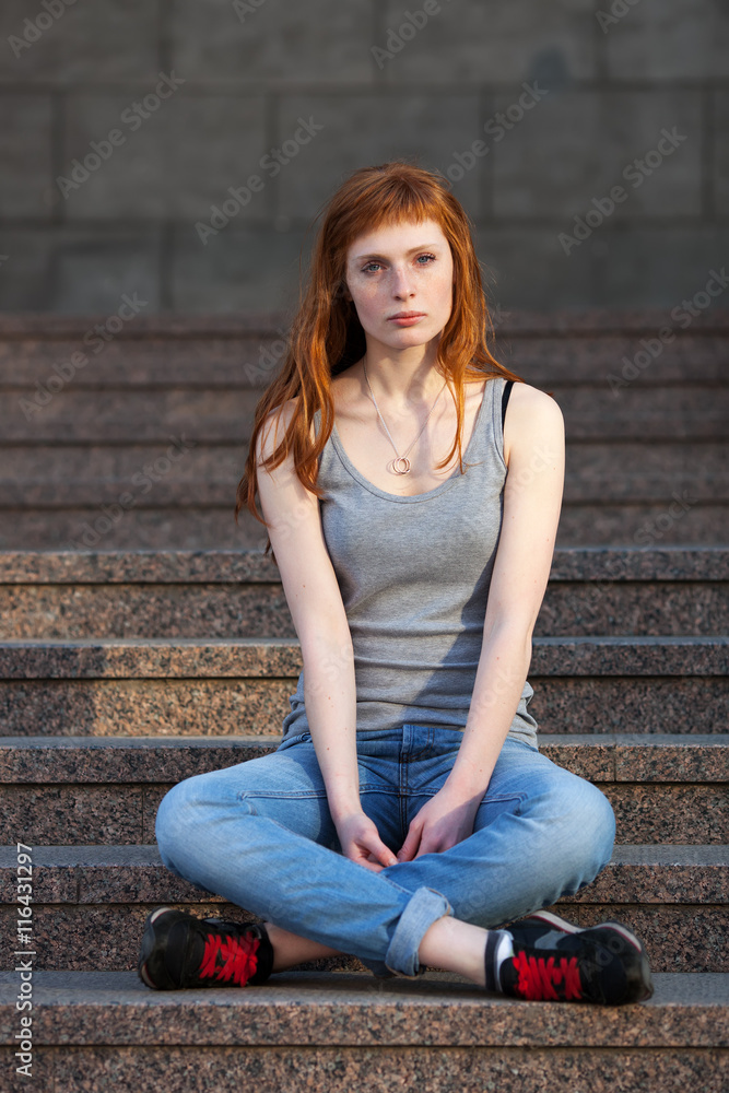 Young redhead caucasian woman in grey shirt blue jeans black sneakers sitting on stairs outdoor portrait Young redhead caucasian woman in grey shirt blue jeans black sneakers sitting on stairs outdoor portrait