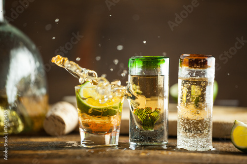 Shot glass of tequila splashing on wood table with lime. Selective focus, blurred background.