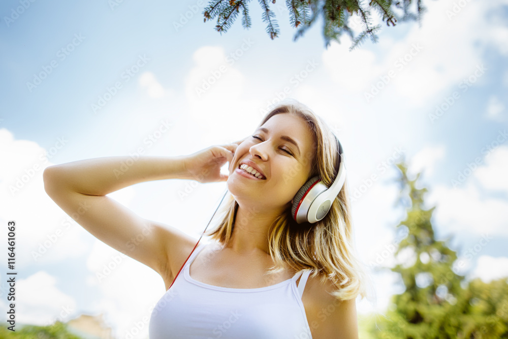 Teenage girl with headphones  outdoors on sunny summer day smiling. Beautiful millennial young woman listening to music relaxing. Horizontal, retouched,