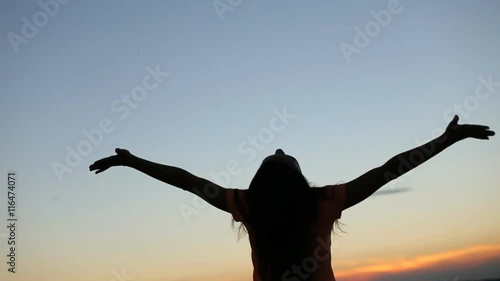 Silhouette of young woman raising hands to sun at sunset. Moment of freedom and happiness. Young woman meditates on the bridge with city landscape background