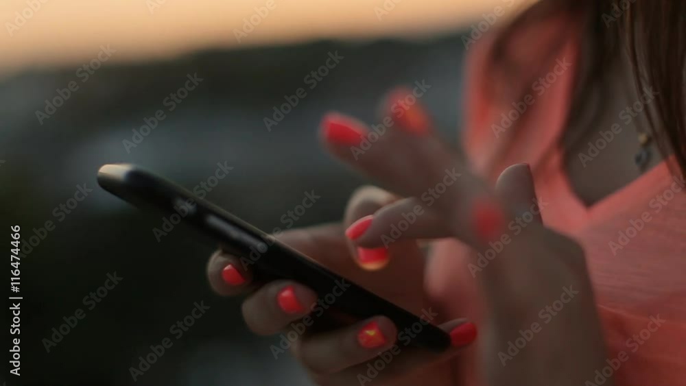 Young woman with nice orange manicure use her mobile phone scrolling outdoor at sunset.Close up. Young woman with nice orange manicure use her mobile phone scrolling outdoor at sunset.Close up.