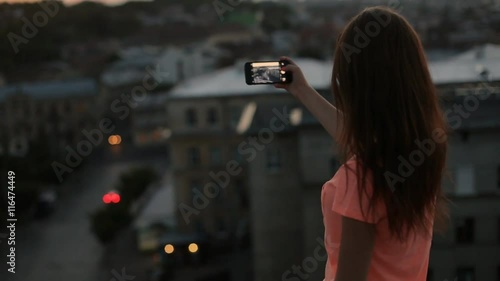 Young pretty woman with long hair taking photo, filming with cell phone at the top of building with city landscape at sunset. View from behind