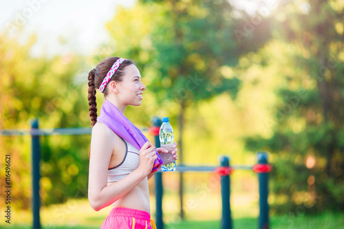 Young girl with botttle of water after running outside. Female fitness model training outside in the park. Concept of healthy and proper nutrition.