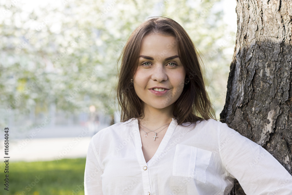 Young beautiful woman in a white shirt standing by the tree in a park smiling dark hair Caucasian Young beautiful woman in a white shirt standing by the tree in a park smiling dark hair Caucasian
