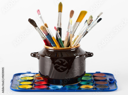 Paint brushes in clay jug and water paints on white background