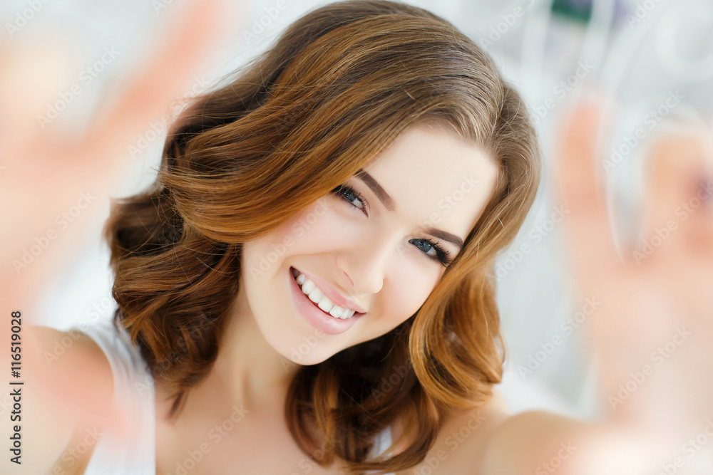 Fototapeta premium Young beautiful woman with wavy red hair and gray eyes,light makeup and cute smile,dressed in a white shirt sitting on white bed in the bedroom,with my hands in front of him,imitates selfie