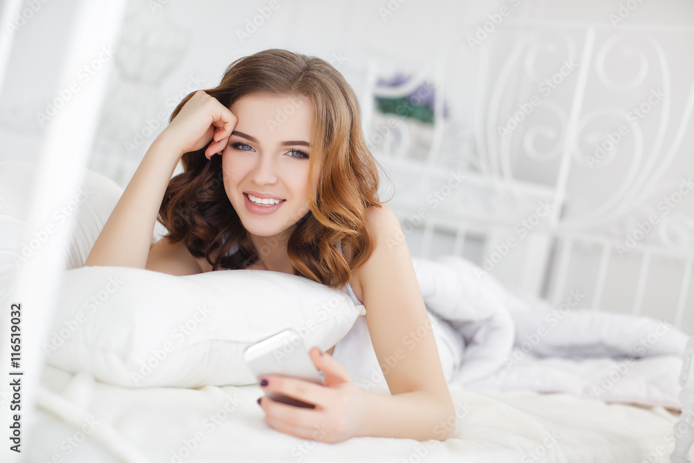 Beautiful girl with wavy red hair and gray eyes,cute smile,dressed in a white t-shirt,waking up in the morning on a white bed in the bedroom,reading a message on a white smartphone Beautiful girl with wavy red hair and gray eyes,cute smile,dressed in a white t-shirt,waking up in the morning on a white bed in the bedroom,reading a message on a white smartphone