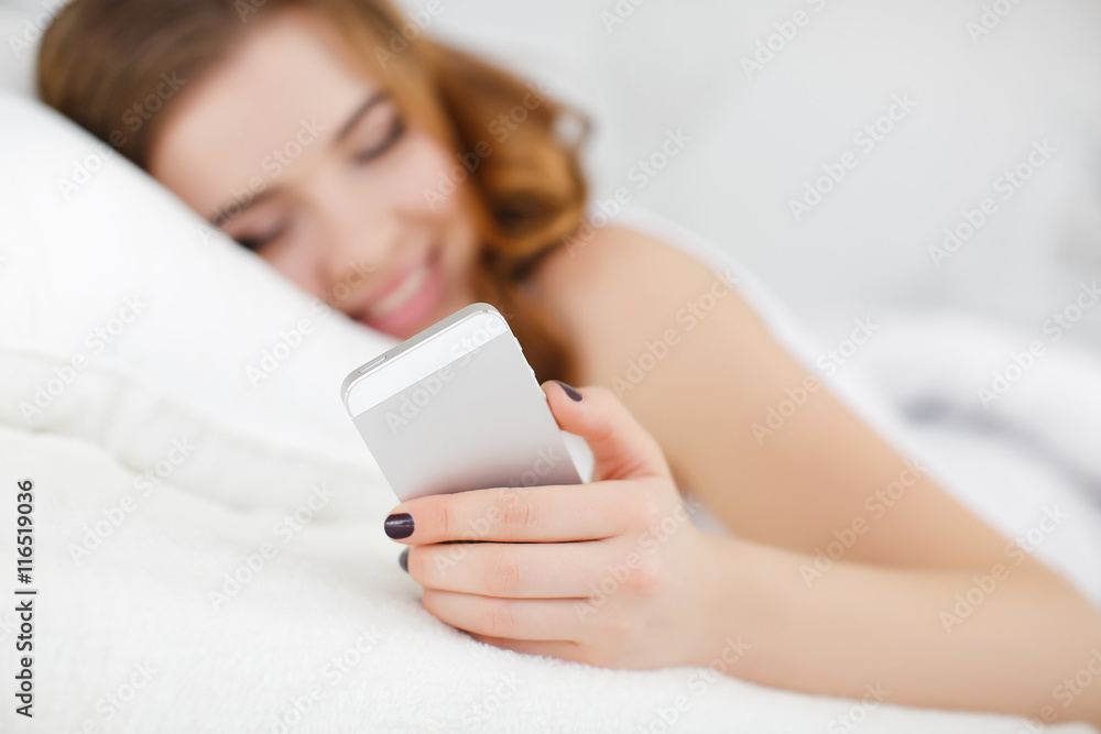 Beautiful girl with wavy red hair and gray eyes,cute smile,dressed in a white t-shirt,waking up in the morning on a white bed in the bedroom,reading a message on a white smartphone Beautiful girl with wavy red hair and gray eyes,cute smile,dressed in a white t-shirt,waking up in the morning on a white bed in the bedroom,reading a message on a white smartphone