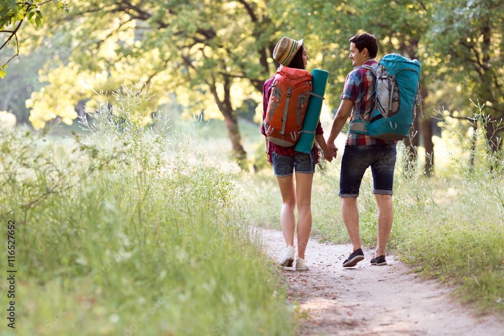 Sporty Family Man and Woman with Backpacks walking along Forest Trail holding Hands with Sunny Background Sporty Family Man and Woman with Backpacks walking along Forest Trail holding Hands with Sunny Background